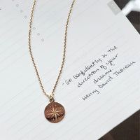 Rose gold necklace with a compass pendant on a white paper background with text.