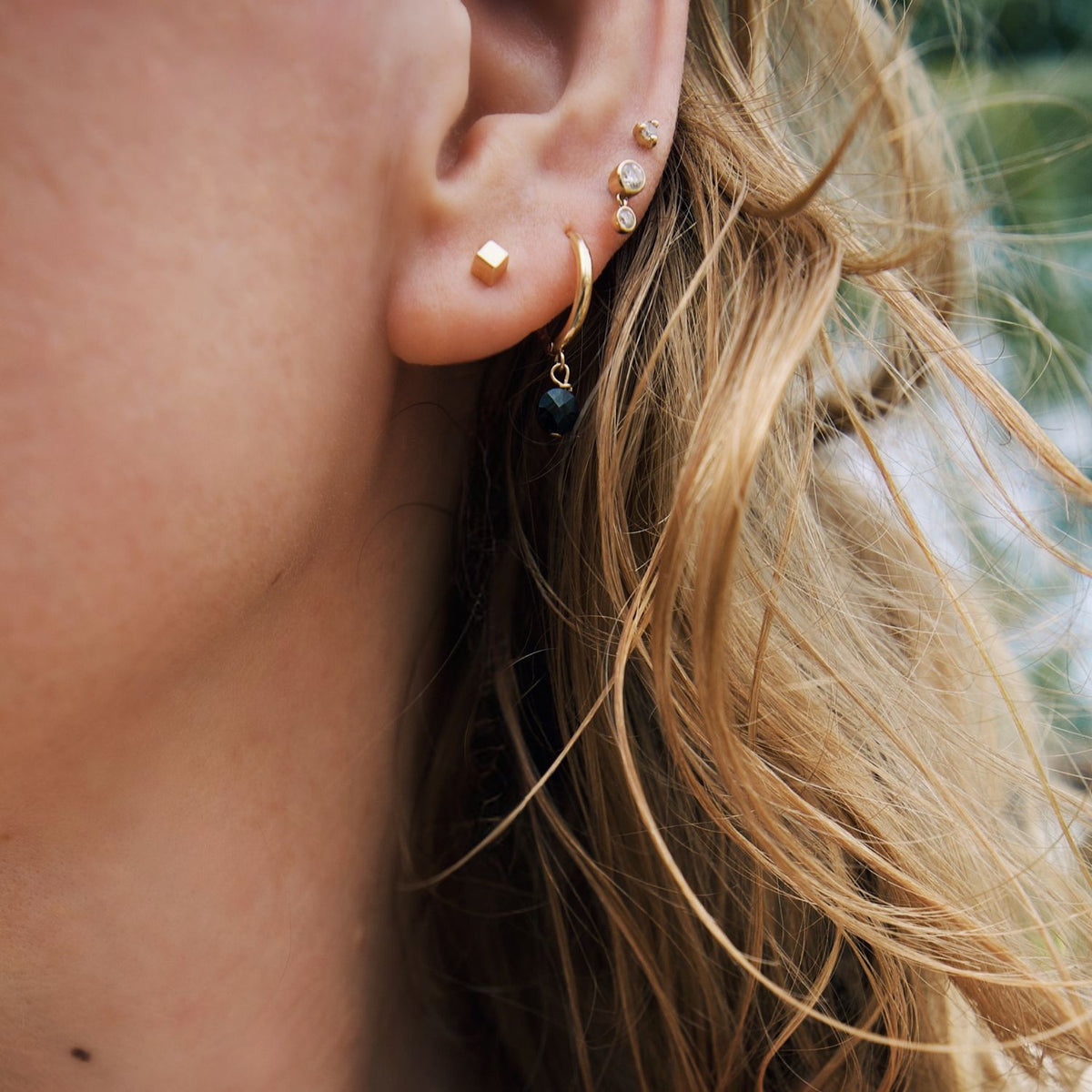 Close-up of an ear wearing gold cube stud earrings with a blurred natural background