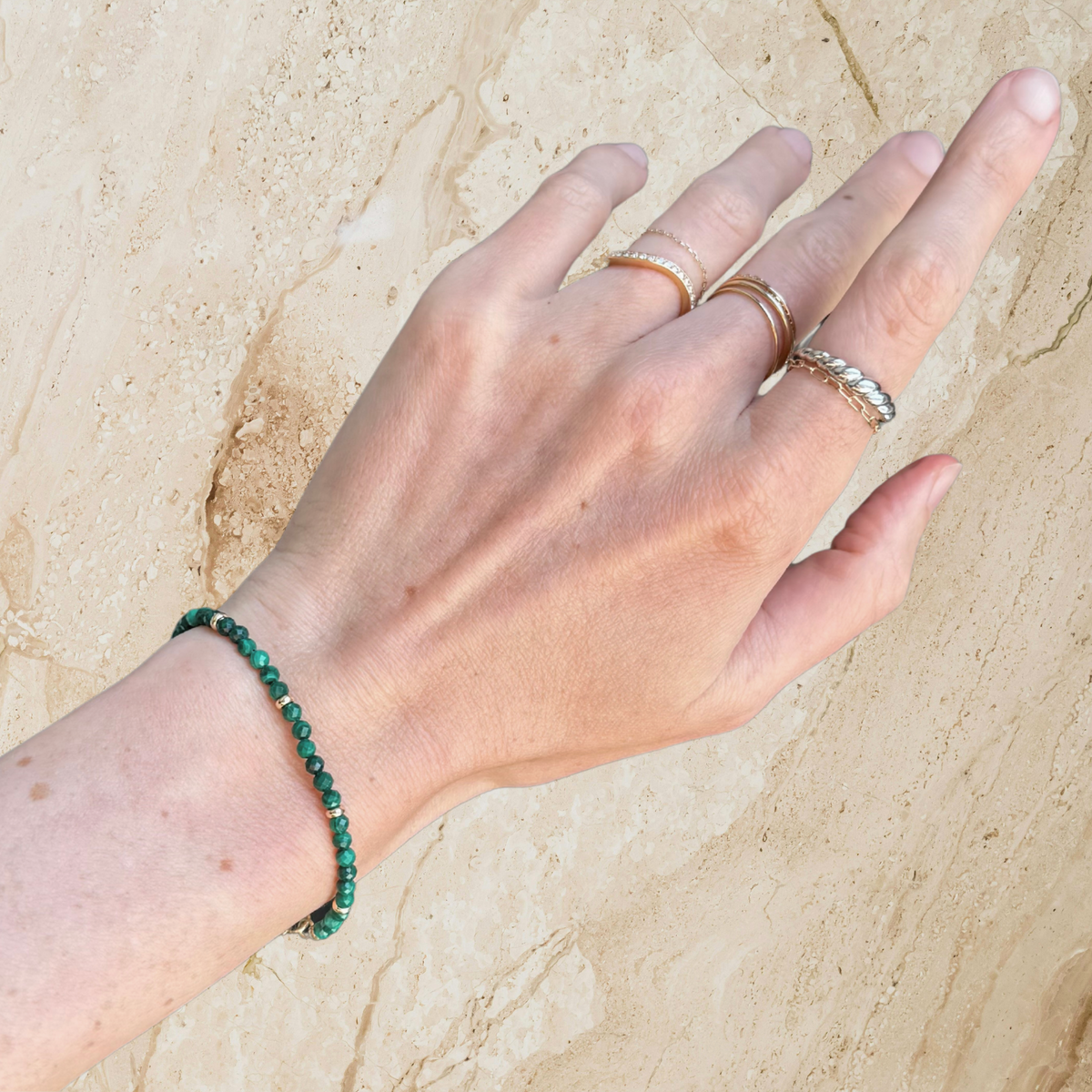 Hand wearing a green malachite beaded bracelet and multiple gold rings on a beige stone background