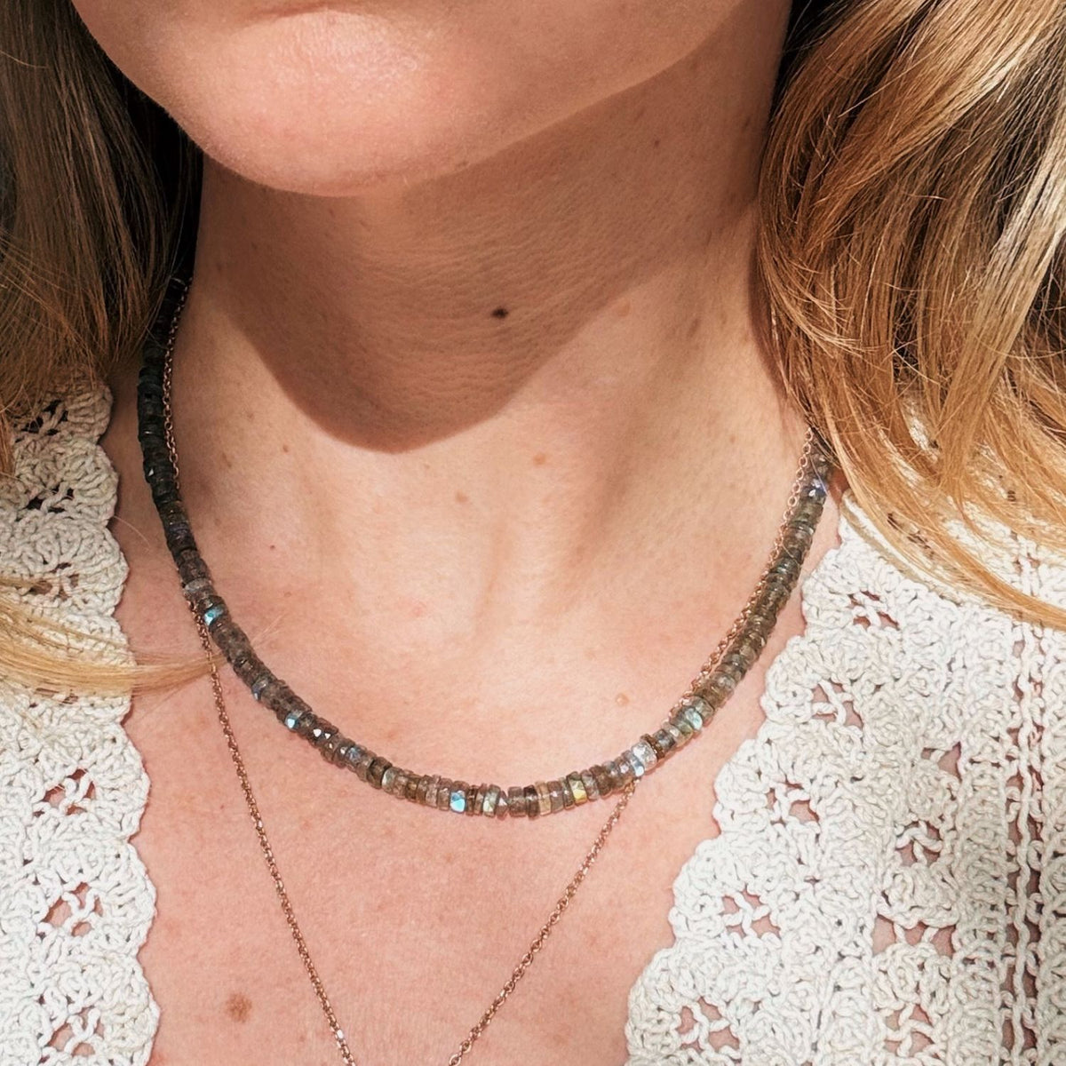 A close-up of a woman wearing a beaded labradorite necklace with faceted gray and iridescent blue-green beads, layered over a delicate gold chain necklace, against a white lace top.