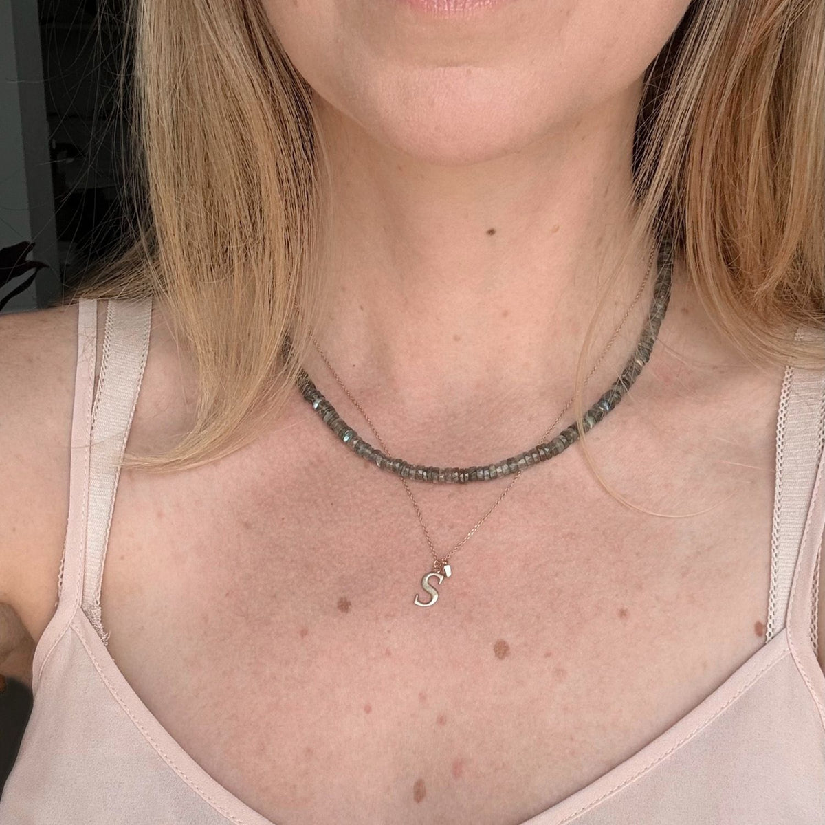 A close-up of a woman wearing a layered necklace set. The top layer is a chunky dark gray labradorite beaded choker, and the bottom layer is a delicate silver chain featuring a small gold 'S' initial pendant.