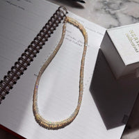 An opal beaded necklace with iridescent, shimmering beads in multicolor tones, laid on an open spiral notebook next to a white gift box.