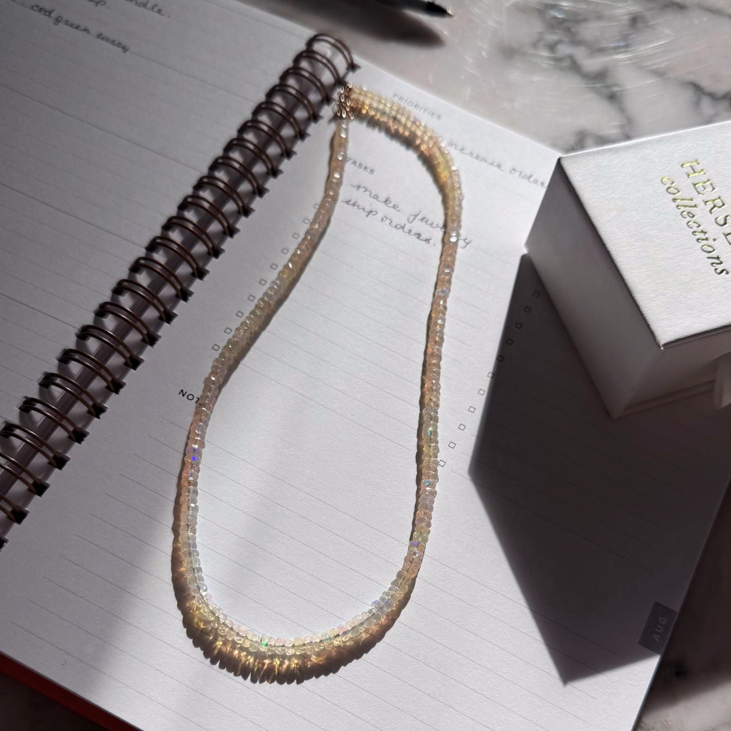 An opal beaded necklace with iridescent, shimmering beads in multicolor tones, laid on an open spiral notebook next to a white gift box.