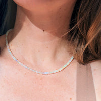 A close-up of a model's neck and collarbone wearing a delicate, sparkling iridescent opal tennis necklace.