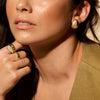Close-up of a woman wearing 14k yellow gold earrings and rings, with a neutral background.