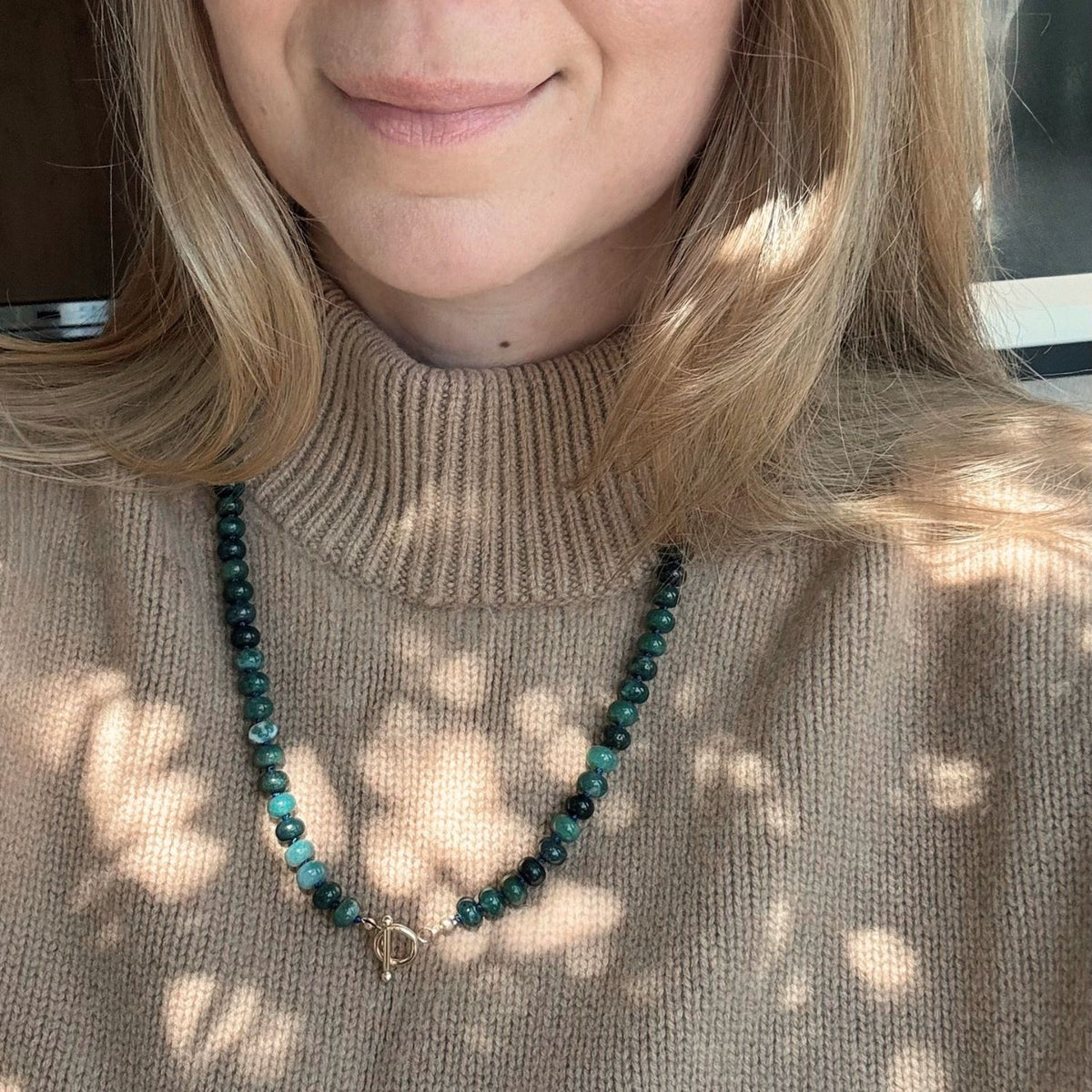 Woman wearing a green bead necklace over a camel colored sweater. 
