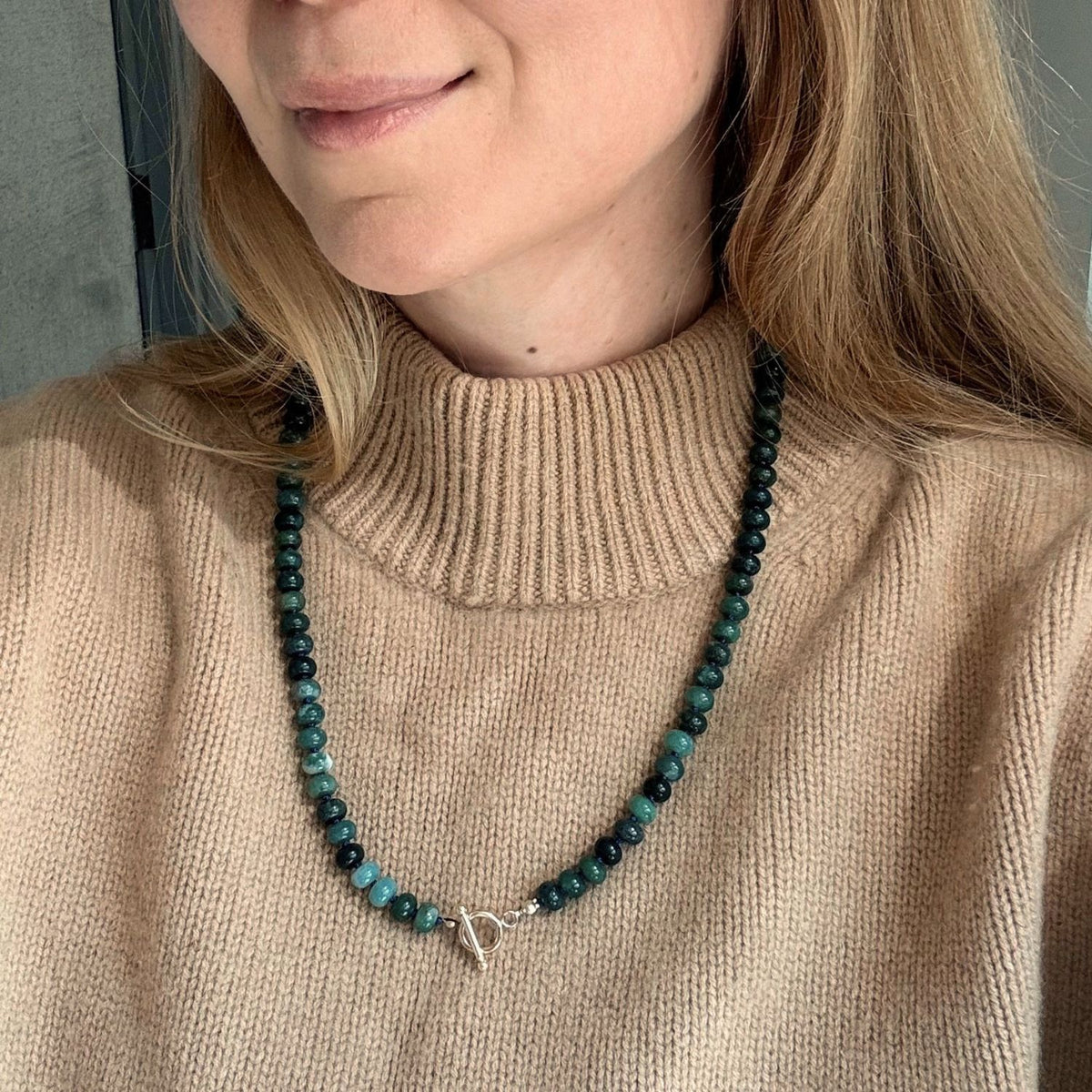 Woman wearing a bold green necklace with a toggle clasp over a tan sweater. 