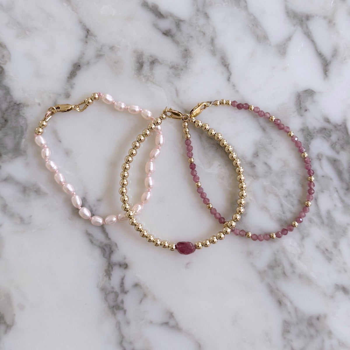 Two pink tourmaline and gold beaded bracelets and one pink pearl bracelet on a marble background