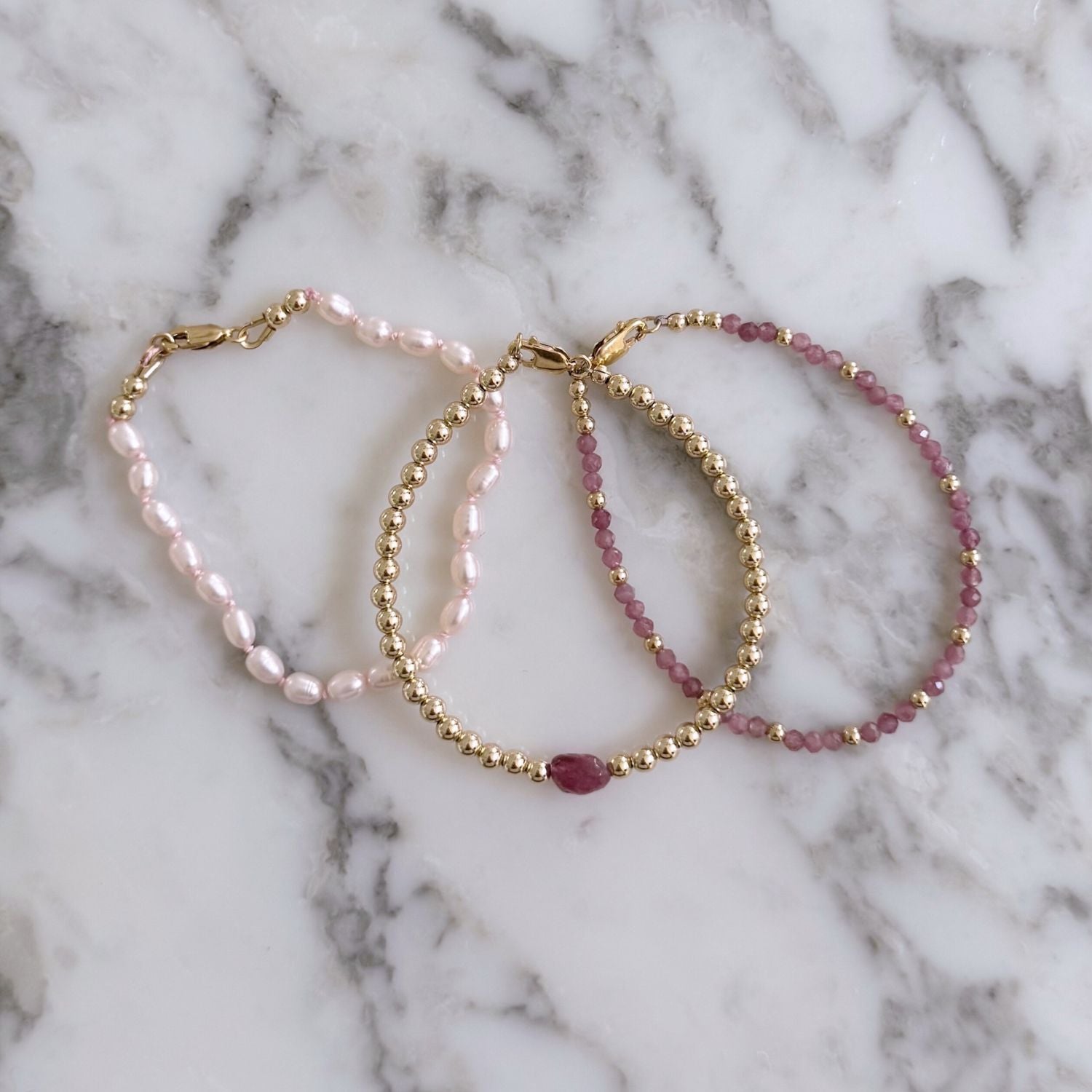 Two pink tourmaline and gold beaded bracelets and one pink pearl bracelet on a marble background