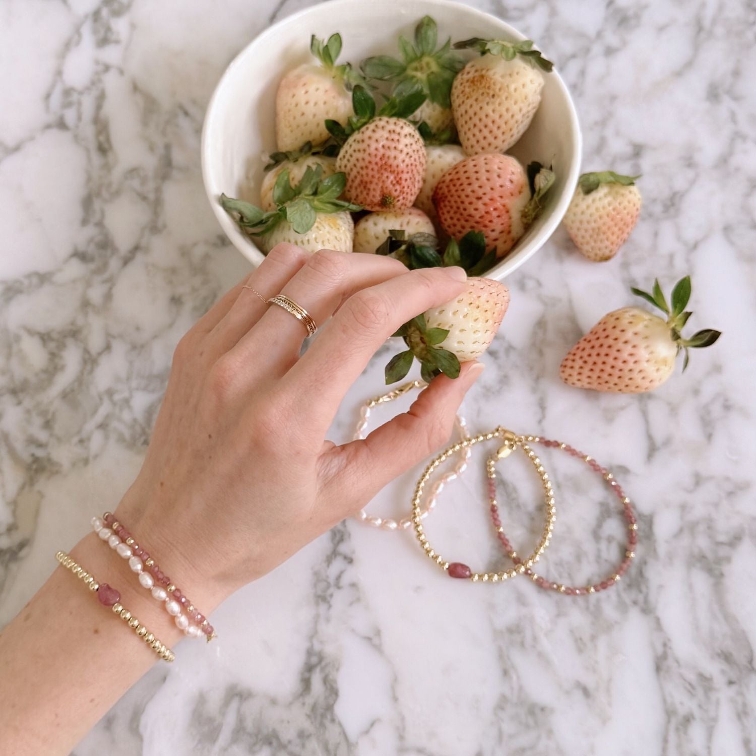 Hand holding a strawberry with a bowl of strawberries on a marble surface showcasing pink and gold bracelets