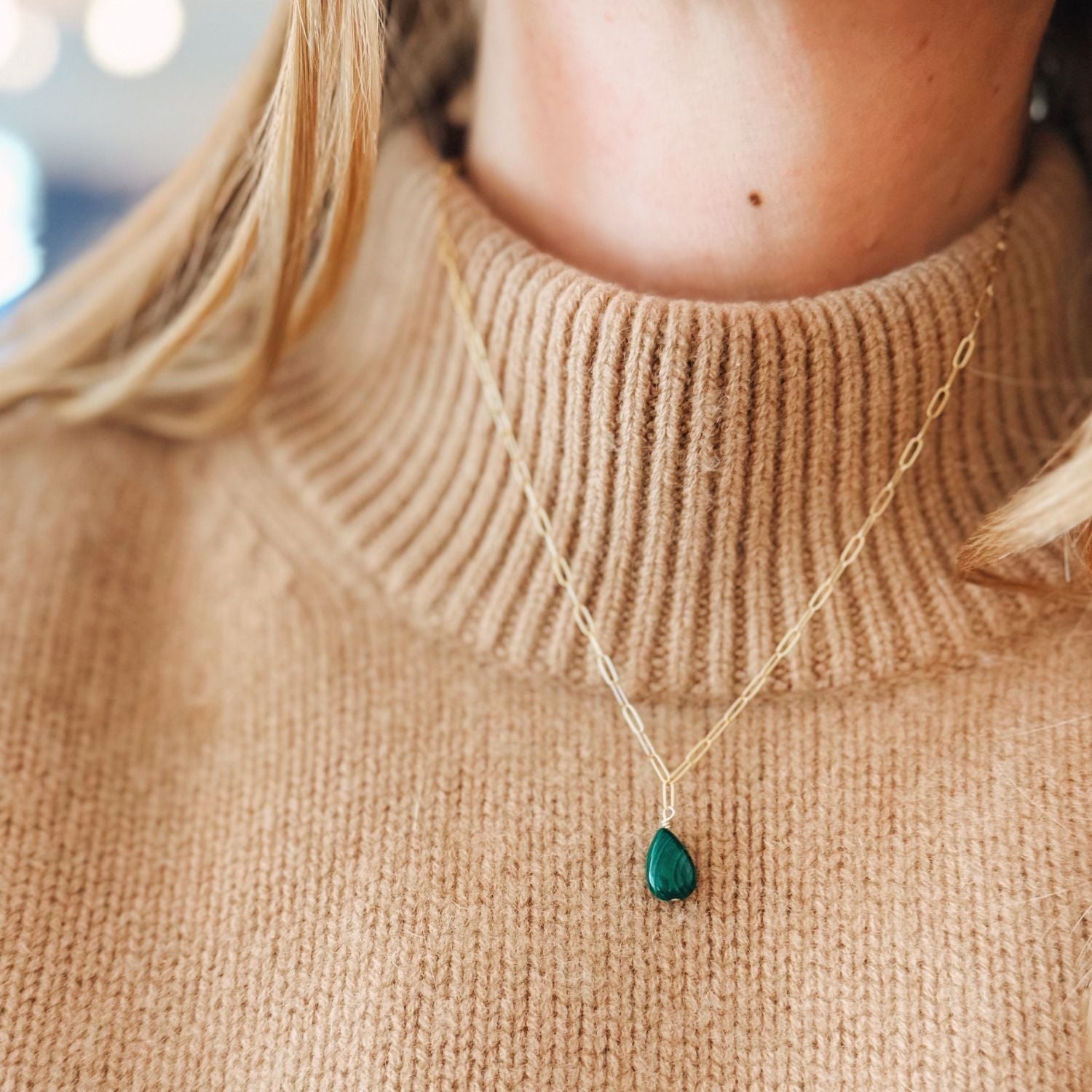 Person wearing a beige sweater with a gold necklace featuring a green teardrop pendant.