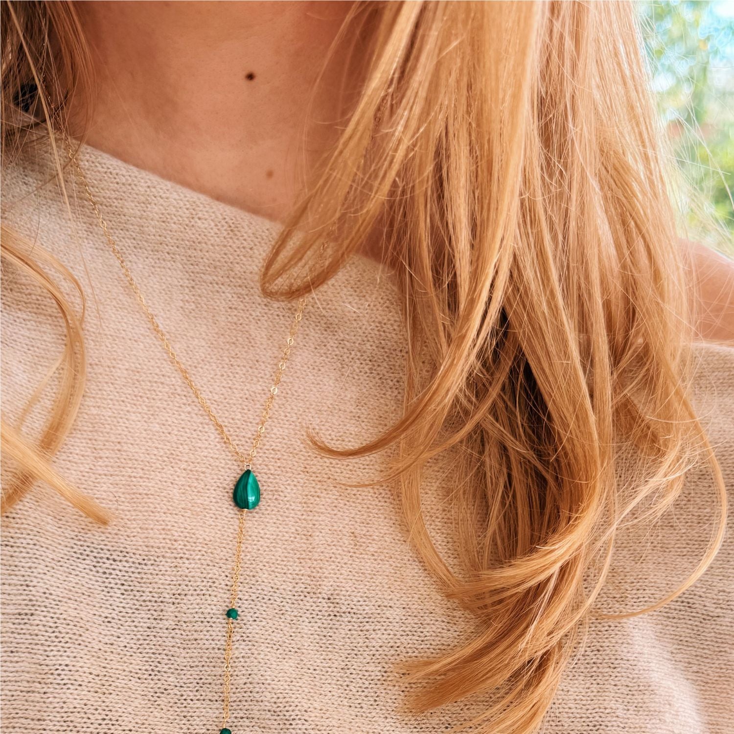 Close-up of a person wearing a gold necklace with a green pendant, blurred background