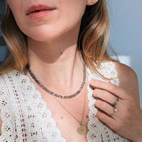 A close-up lifestyle shot of a woman wearing layered necklaces. She is wearing a short, textured labradorite beaded necklace, a delicate gold chain with a round gold sunburst pendant, and a silver ring on her finger.