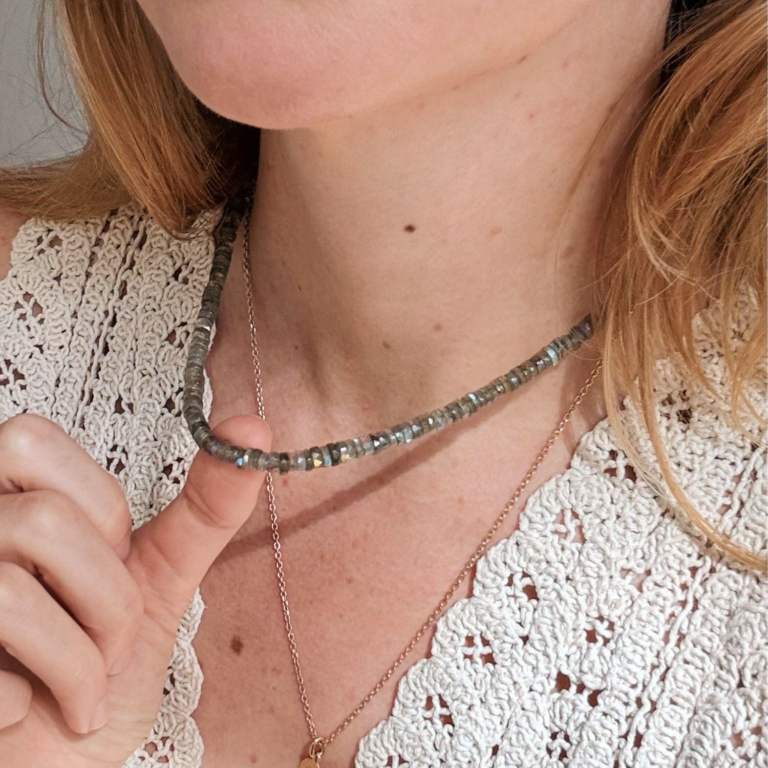 A close-up of a woman wearing a beaded labradorite choker necklace made of gray and green faceted beads, layered with a delicate gold chain necklace. She is wearing a white crochet top.
