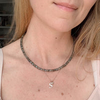 Close-up of a woman wearing a layered necklace set. The top layer is a chunky beaded choker in gray and iridescent stones, and the bottom layer is a delicate silver chain with a small gold initial 'S' pendant.