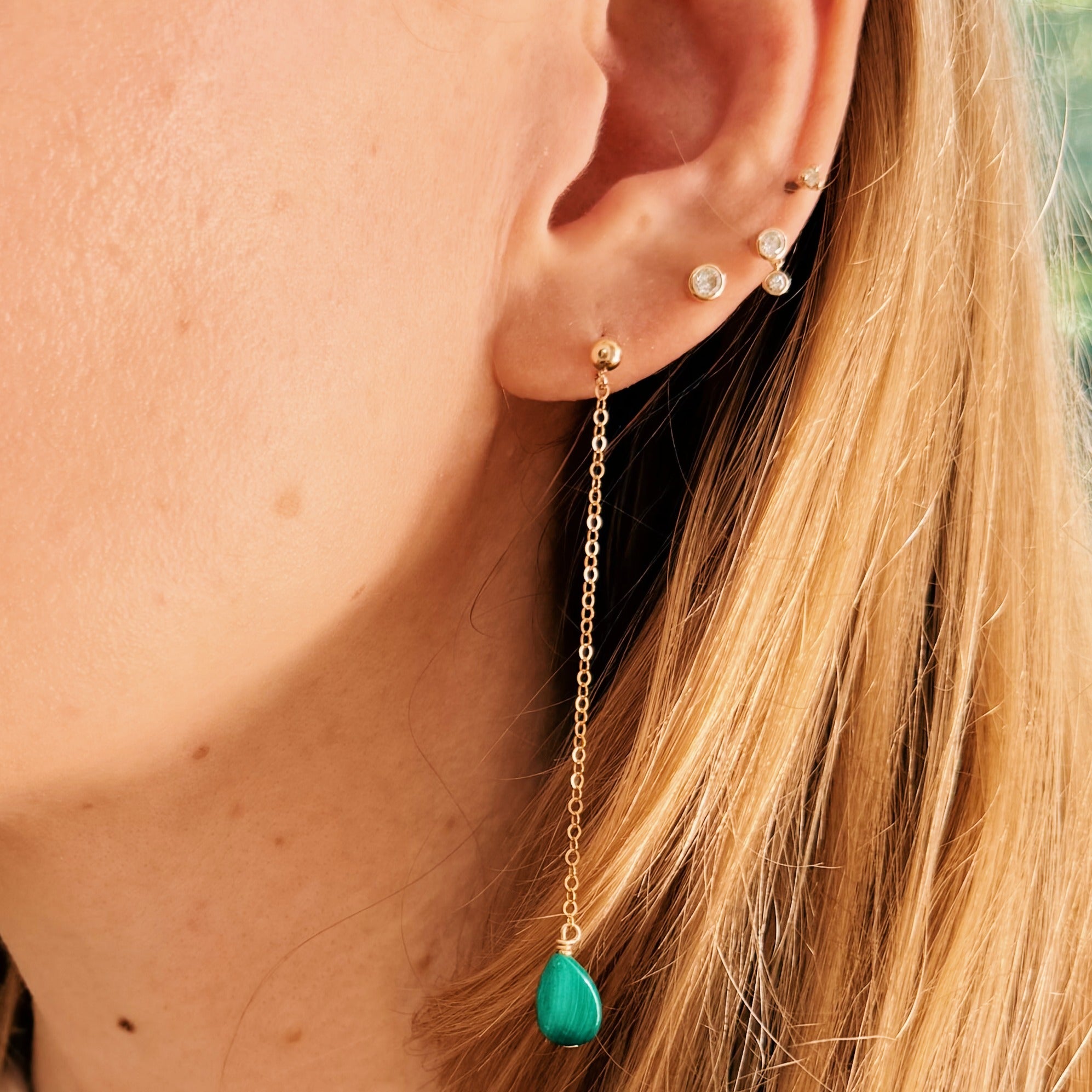 Close-up of an ear with gold chain earrings featuring a green teardrop pendant.