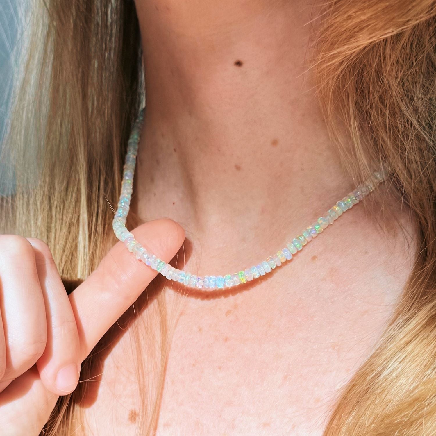 A close-up of a delicate beaded necklace worn on a person's neck, with a finger pointing to the front of the necklace. The opal beads are iridescent and reflect pastel colors including white, light blue, and hints of green and yellow.